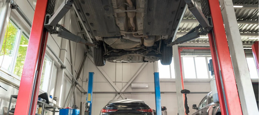 A vehicle raised on a red hydraulic lift in an auto repair garage, displaying its undercarriage with other workshop equipment visible in the background.