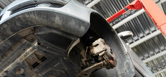 A close-up view of the underside of a lifted vehicle in a workshop, showing the exposed suspension and brake components with a red two-post lift in the background.