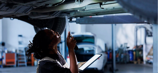 A mechanic holding a flashlight and tablet inspects the underside of a vehicle lifted on a car lift in a brightly lit auto repair shop.