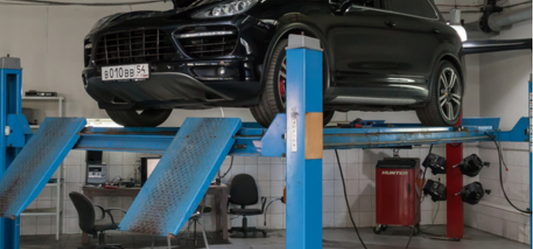 Vehicle lifted on a four-post lift equipped with a rolling jack in a professional auto repair shop, showing undercarriage access for maintenance and wheel alignment tasks.